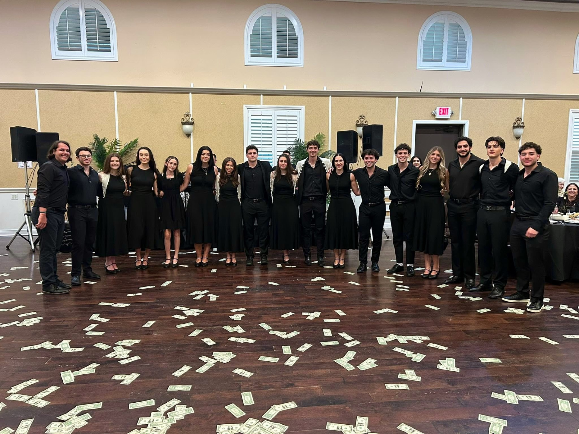 Young members in black attire posing arm in arm on a banquet dance floor with money scattered in front